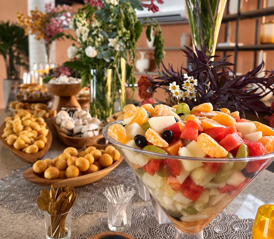 Mesa de brunch com salada de frutas em taça, salgados assados e arranjos de flores.