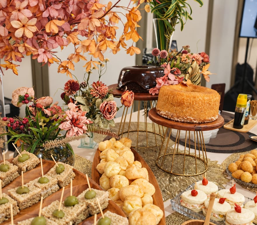 Mesa de coffee break com bolo, pães de queijo, canapés e docinhos entre arranjos de flores.